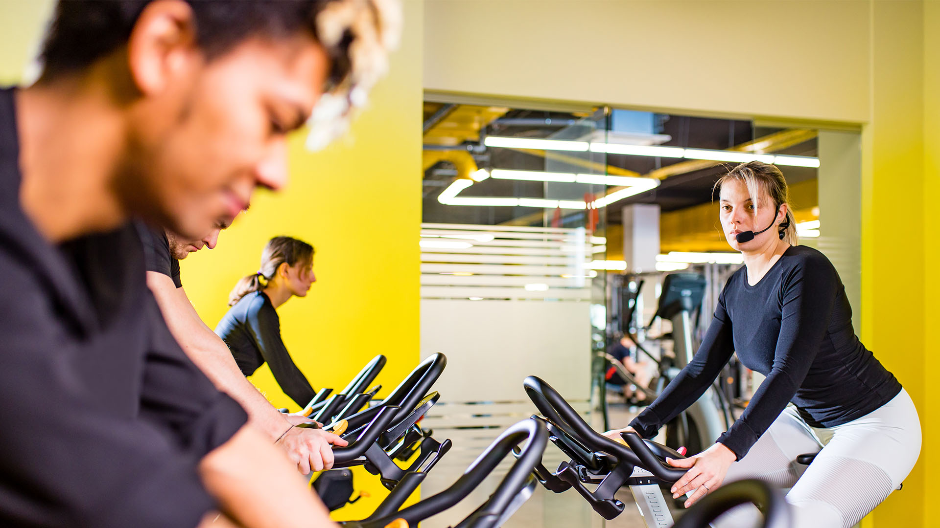 People in a fitness class are riding stationary bikes; the instructor, wearing a headset, offers tips and tricks from the front. The well-lit room boasts modern decor, creating the perfect gym environment.