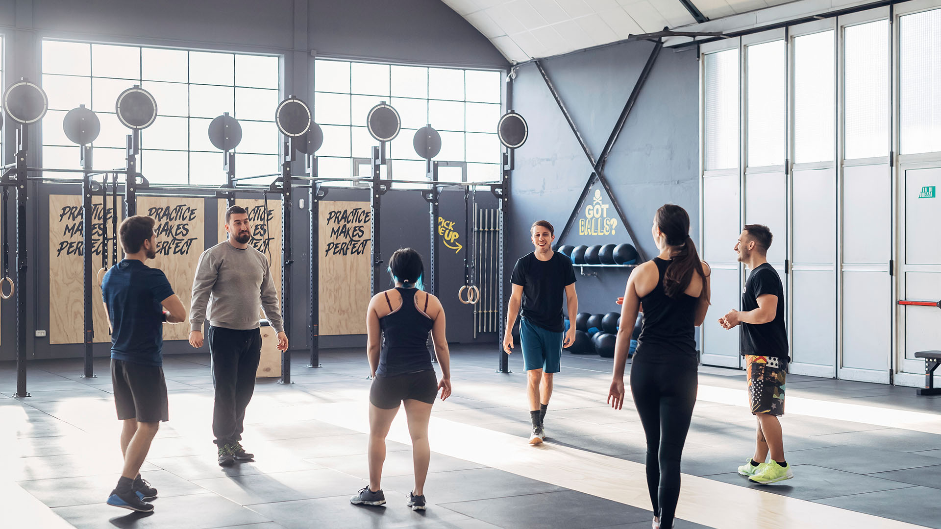 A group of people stands in a gym, engaging in conversation. The gym features equipment such as pull-up bars and stacked weight plates. Sunlight streams through large windows, creating an inviting atmosphere. The wall reads "Practice makes perfect," offering tips and tricks for finding the perfect gym.