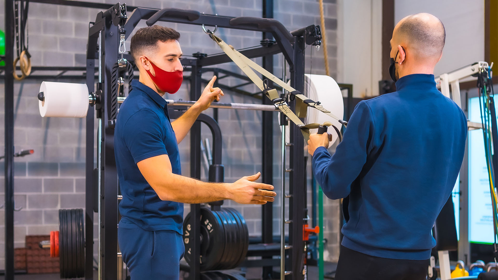 Two men, one wearing a red mask and the other a black mask, are using resistance bands in Perfect Gym, surrounded by various exercise equipment.