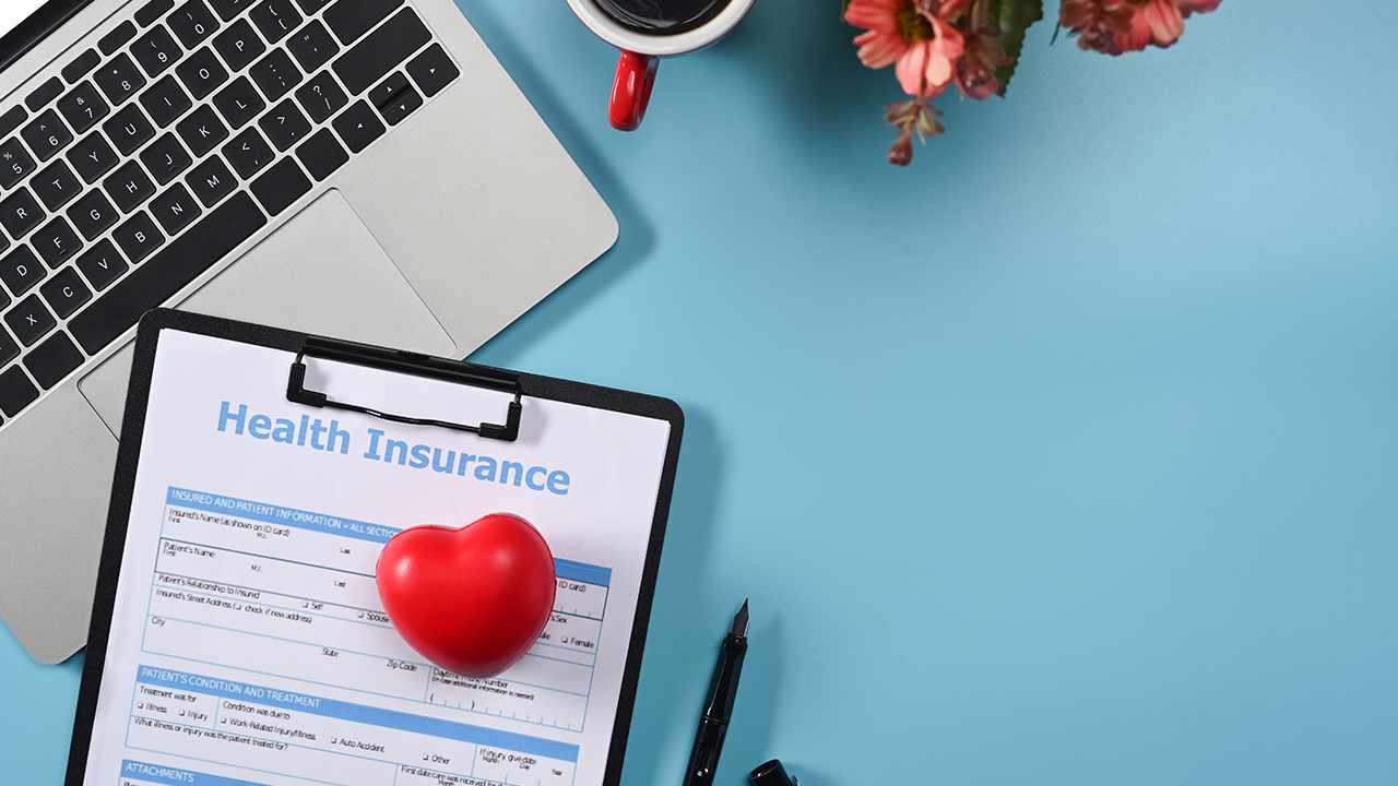 A health insurance form on a clipboard lies on a desk with a red heart-shaped object, pen, laptop, coffee cup, and flowers. Understanding Insurance Basics is essential to ensure adequate Health Coverage for your needs.