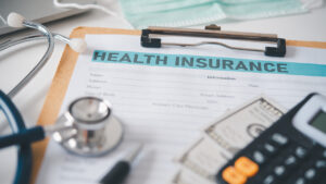 A health insurance form on a clipboard, accompanied by a stethoscope, a calculator, a pen, money, and a face mask nearby—covering all the basics.